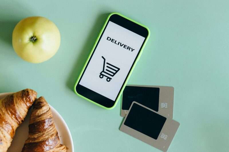 smartfon z napisem "DELIVERY" na ekranie, jabłko, rogaliki i karty płatnicze leżące na stole