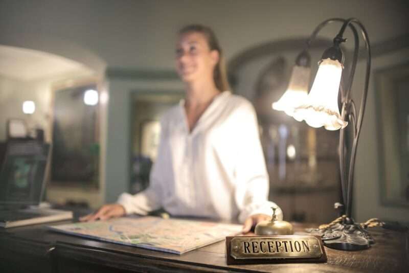 Recepcjonistka przy hotelowym biurku z mapą, dzwonkiem i lampą.