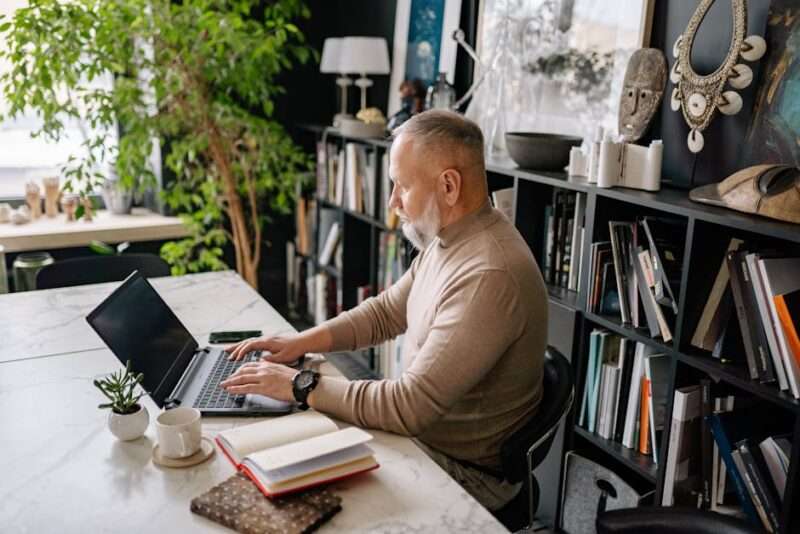 Starszy mężczyzna z brodą pracuje na laptopie przy biurku z książkami.