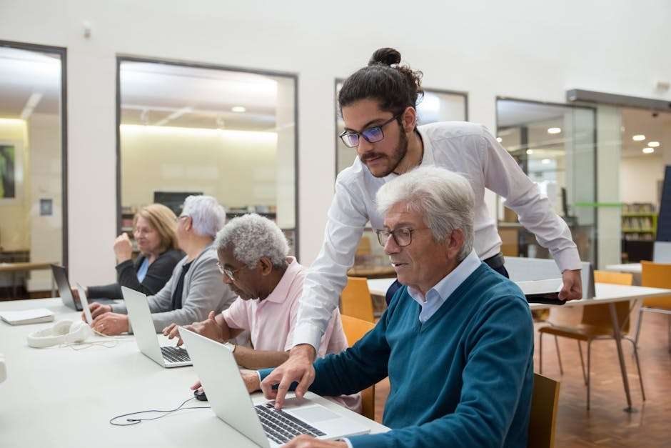 Młody mężczyzna pomaga starszemu mężczyźnie w obsłudze laptopa przy biurku w bibliotece.