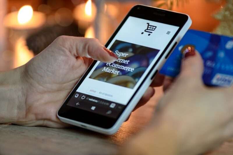 Dłonie trzymają smartfon z otwartą stroną sklepu internetowego oraz kartę płatniczą.