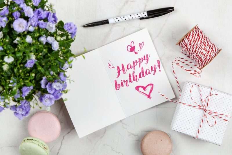 otwarty zeszyt z zapisanymi słowami "Happy birthday", kwiaty, prezenty i długopis leżące na stole